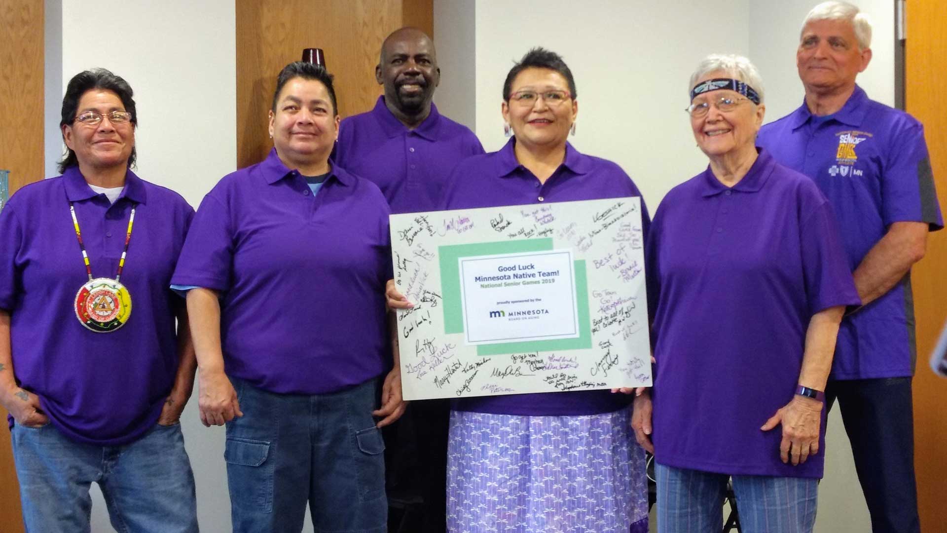 A picture of 6 people standing together in purple shirts. The person in the middle is holding a sign that reads "Good luck Minnesota Native Team! National Senior Games 2019." The sign has many handwritten notes on it.