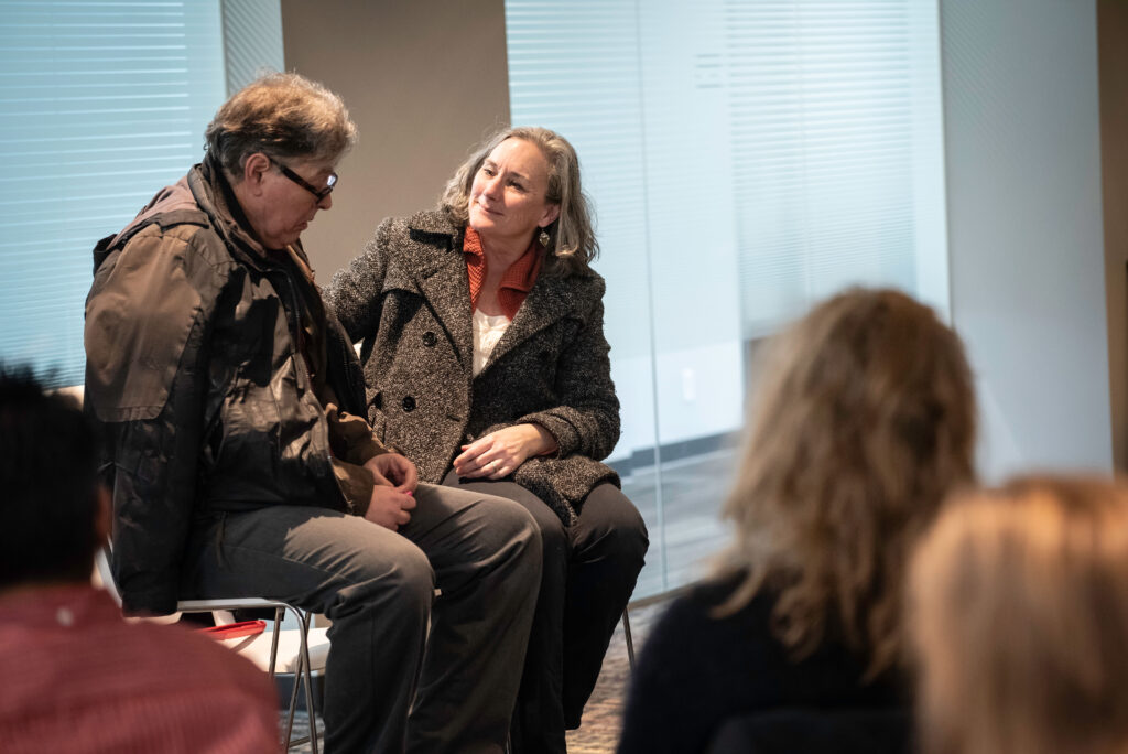 A photo of two people seated, talking with one another as part of a play performance.