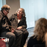 A photo of two people seated, talking with one another as part of a play performance.