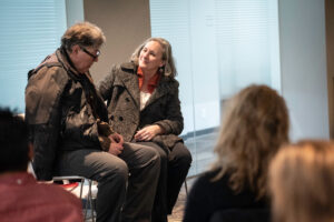 A photo of two people seated, talking with one another as part of a play performance.