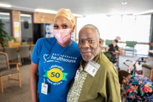 Two people posing together indoors. The person on the left is wearing a facemask.