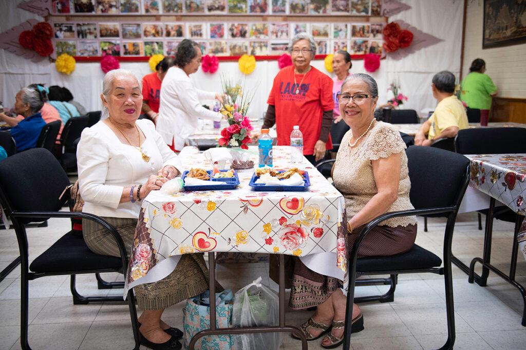 A photo of two women eating together. Behind them are other people eating together at several tables.