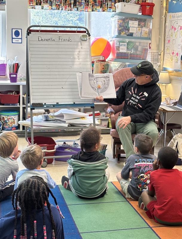 Photo of a person reading a book to a classroom of elementary students seated on the floor.