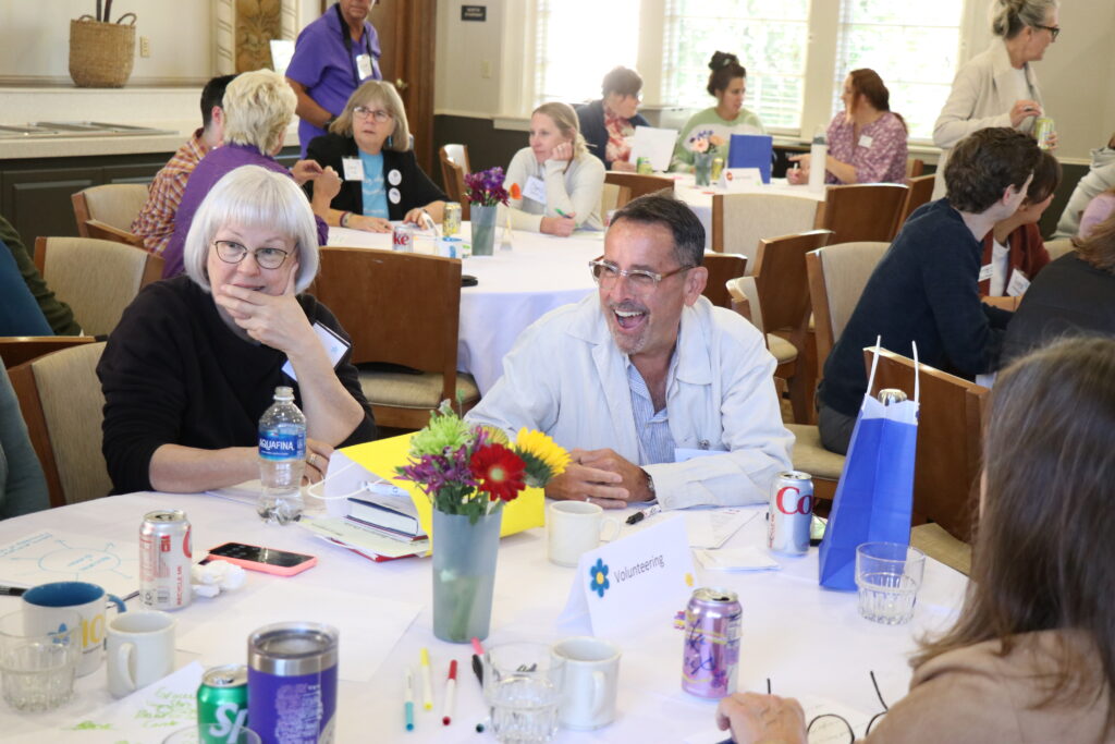 Photo of three people sitting at a table talking in a conference room full of people.