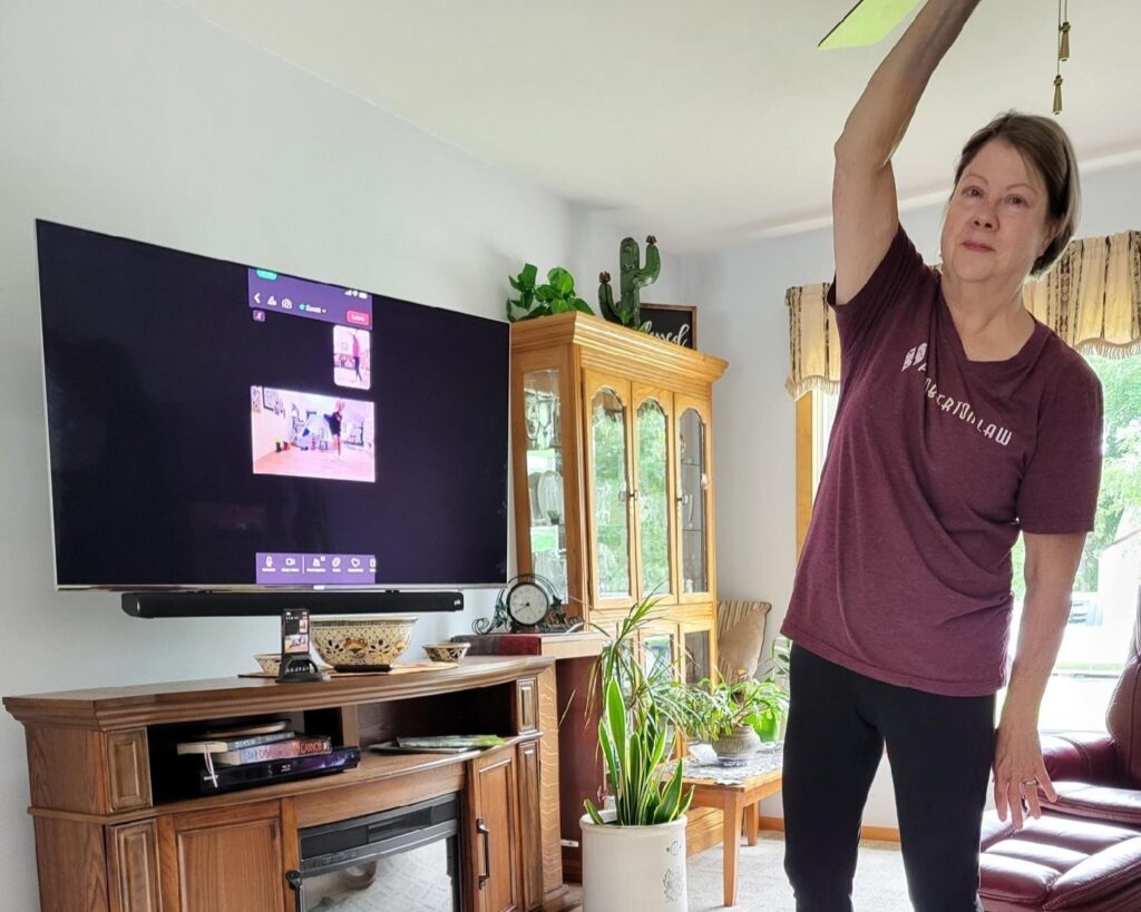 Senior woman Karel is stretching right arm above her head as part of a video exercise class.