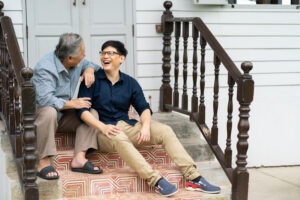 Photo of an older adult and a younger man sitting on the front steps of a house, laughing together.