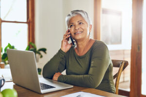 Photo of an older adult speaking on a phone while sitting at a desk with a laptop in front of her.